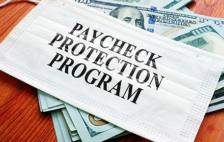 White Facemask Over Dollar Bills With the Words Paycheck Protection Program